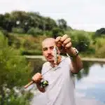 Fisherman proudly displays his catch, a small fish, while holding a fishing rod by a tranquil lake surrounded by lush green trees.