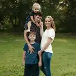 A family of four stands in a grassy field: a man with a toddler on his shoulders, a young boy in front, and a woman beside him. They are posing for a photo, creating a warm, family moment.